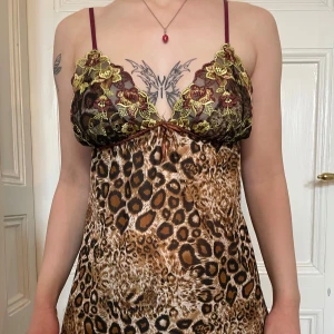 Leopardmönstrad nattlinne - Säljer en kort klänning med leopardmönster och tunna axelband. Överdelen har broderad spets i gult och vinrött med blommor och v-ringning. Kjolen är glansig med volangkant nertill. Perfekt för dig som vill sticka ut med en unik look.