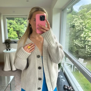 Beige kofta Kashmir  - Supermjuk och oversized beige kofta med stora knappar och bred ribbad kant. Tillverkad i en lyxig blandning av ull och kashmir, perfekt för lager-på-lager. Djup v-ringning och långa ärmar ger en chill och avslappnad vibe.