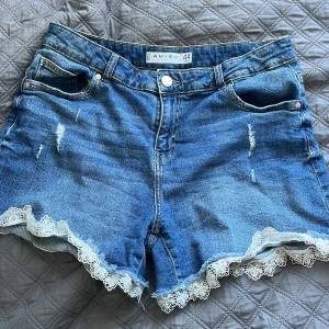 Blå jeansshorts med spets från Amisu - Snygga blå jeansshorts från Amisu med ljusa slitningar och vit spetskant längst ner. Klassisk femficksmodell med knapp och dragkedja framtill. Perfekta för sommaren när du vill ha en chill men ändå söt look.