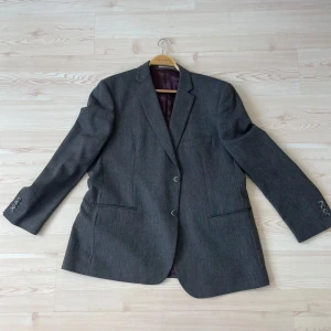 Grå kavaj från Dressmann, stl 58 - Snygg grå kavaj från Dressmann i klassisk modell med två knappar framtill och flera innerfickor. Fodret är blått och vinrött med bruna detaljer. Perfekt för dig som vill ha en stilren och tidlös look.