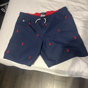 Marinblå badshorts Polo Ralph Lauren - Snygga marinblå badshorts från Polo Ralph Lauren med röd broderad logga över hela tyget. Shortsen har elastisk midja med vit snörning och röd insida. Perfekta för stranden eller poolen, klassisk stil och bekväm passform.