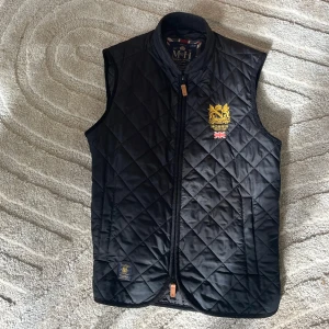 Svart quiltad väst från Morris - Svart quiltad väst från Morris med guldfärgat broderat emblem och brittisk flagga på bröstet. Västen har dragkedja framtill och två fickor. Snygg och stilren design med diskret logga nertill. Perfekt för lager-på-lager och klassisk look.