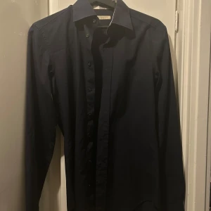 Mörkblå skjorta från Burberry - Snygg mörkblå skjorta från Burberry med klassiska rutiga detaljer på insidan av kragen och ärmsluten. Skjortan har knappar framtill och är långärmad. Perfekt för dig som gillar stilrena plagg med en twist.