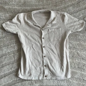 Ljusbeige kortärmad skjorta med knappar - Snygg ljusbeige skjorta med korta ärmar och klassisk krage. Skjortan har knappar framtill och en bröstficka. Materialet är mjukt och har ett diskret mönster som ger en stilren känsla. Perfekt för en chill och avslappnad look.