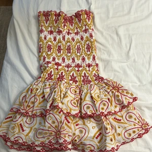 Vit och röd kortklänning Design by Amor - Supersöt kortklänning från Design by Amor med smockad överdel och volangig kjol. Klänningen är vit med broderade mönster i rött och gult, och har fina detaljer längs kanterna. Perfekt för sommarens alla tillfällen!