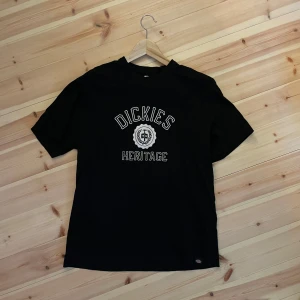 Svart Dickies Heritage t-shirt - Svart t-shirt från Dickies med trycket 'Dickies Heritage' och emblem framtill. Klassisk passform med korta ärmar och rund hals. Perfekt för dig som gillar streetwear och vill ha en stilren look med tydlig logga. Storlek är S men passar som M