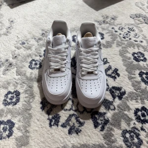 Nike Air Force 1 helvita sneakers - Klassiska Nike Air Force 1 i helvitt skinn med perforerad tå, rund tå och platt sula. Snygg och tidlös modell med snörning och diskret Nike-logga på sidan. Perfekta för dig som gillar cleana och stilrena sneakers.