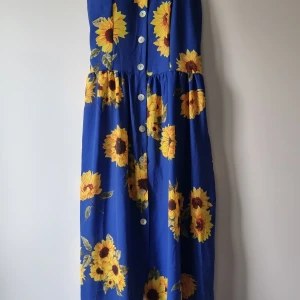 Blå långklänning med solrosor - Blå långklänning med stora gula solrosor och vita knappar framtill. Klänningen har ärmlös design och en luftig passform, perfekt för varma dagar. Tyget känns lätt och somrigt, och mönstret är riktigt färgstarkt och glatt.
