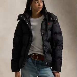 Svart pufferjacka från Polo Ralph Lauren - Snygg svart pufferjacka från Polo Ralph Lauren med röd logga på bröstet. Jackan har hög krage, avtagbar huva och dragkedja framtill. Fylld för extra värme och har flera praktiska fickor. Perfekt för kalla dagar och riktigt street. använd endast 2 gånger, svårt att hitta en jacka med bättre skick. 