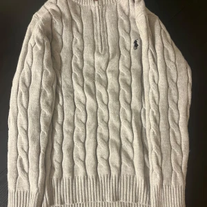 Ljusgrå stickad tröja  - Kabelstickad ljusgrå tröja   med half zip och broderad logga på bröstet. Tröjan har lång ärm, ribbade muddar och är tillverkad i mjukt stickat material. Perfekt för dig som gillar klassisk och stilren look.  OBS!! Inte äkta