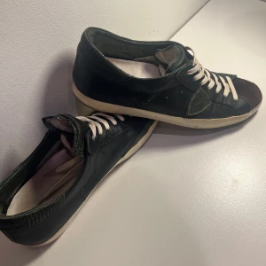 Svarta sneakers från Philippe Model - Snygga svarta sneakers från Philippe Model med vita snören, mörkgrön häl och detaljer samt mocka på tån. Skorna har en vit sula och är tillverkade i skinn och mocka för en cool och urban look. Säljes för en billig peng då jag ej använder dem! Bara att skriva om du har frågor. /