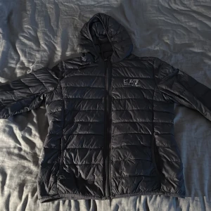 Svart dunjacka från EA7 Emporio Armani - Mörk grå dunjacka från EA7 Emporio Armani med huva och horisontella sömmar. Jackan har dragkedja framtill och logga på bröstet. Perfekt för kyliga dagar och har en clean, sportig look som funkar till det mesta.