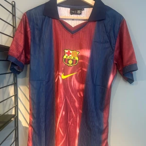 Barça x Cactus Jack fotbollströja Nike - Snygg Barcelona fotbollströja i rödblått med klassiska vertikala ränder, gul Nike-logga och FCB-emblem på bröstet. Baksidan har stort nummer 2, 'Cactus Jack' och 'BARÇA' i vitt. Skön krage och korta ärmar, perfekt för dig som vill sticka ut på planen eller gatan. Helt ny med tagg en replika
