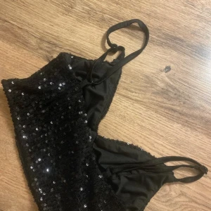 Svart paljett bandeau topp - Snygg svart bandeau topp med tunna axelband och täckt av glittrande paljetter. Perfekt för dig som vill sticka ut och glänsa. Toppens material är mjukt och stretchigt för en bekväm passform.