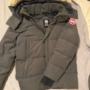 Grå wyndham parka från Canada Goose - Säljer en grå dunjacka från Canada Goose med ikonisk patch på ärmen och stor huva med äkta päls. Jackan har dragkedja, knappar och två stora fickor framtill. Perfekt för kalla vinterdagar och riktigt snygg streetstil. Bra skick, precis kemtvättat den och inte använd den efter.