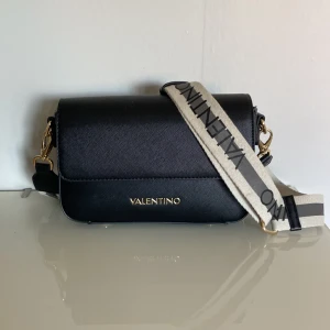 Svart axelväska från Valentino - Snygg svart axelväska från Valentino med guldiga metalldetaljer och avtagbar bred axelrem i beige med svart logga. Väskan är tillverkad i skinnliknande material och har en stilren, rektangulär form. Perfekt för dig som vill ha en trendig och praktisk väska.