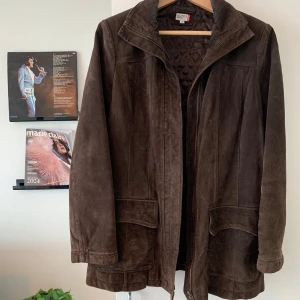 Brun jacka med mockaeffekt - Snygg brun ”mockajacka” från NKD, köpt secondhand med klassisk krage, dragkedja och två stora fickor framtill. Jackan har quiltat innerfoder och raka, långa ärmar. Perfekt för dig som gillar vintagevibbar och tidlös stil.