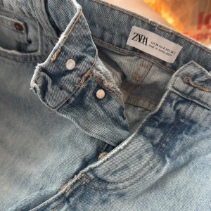 Ljusblå jeans från Zara med rå kant - Säljer ett par ljusblå jeans från Zara med snygg rå kant nertill och klassisk femficksdesign. Jeansen har knäppning framtill och är tillverkade i bomull. Perfekta för en avslappnad och trendig look.