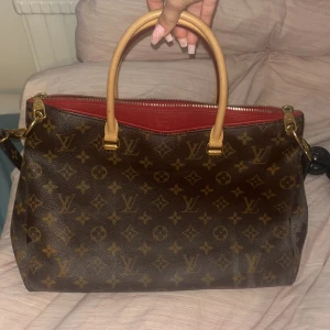Louis Vuitton Monogram Handväska - Säljer en klassisk Louis Vuitton handväska i brunt monogrammönster med guldiga detaljer och röda insidor. Väskan har två rejäla handtag i ljust skinn, dragkedja upptill och rymligt fack inuti. Perfekt för dig som vill ha en ikonisk och lyxig accessoar.
