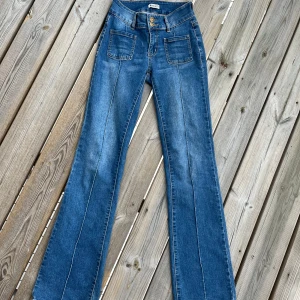 Blå bootcut jeans från Madlady - Snygga blå jeans från Madlady med bootcut-ben och normal midja. Jeansen har framfickor med raka kanter, dubbla knappar i midjan och dekorativa sömmar längs benen. Baktill finns fickor med lock och knapp. Perfekta för en trendig och avslappnad look.  Storlek 32/regular längd.