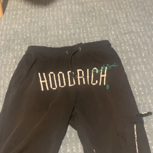 Svarta mjukisbyxor från Hoodrich - Svarta mjukisbyxor från Hoodrich med stor vit logga framtill och broderad text på benet. Byxorna har snörning i midjan, muddar vid bensluten och en ficka med dragkedja på sidan. Perfekta för chill eller streetwear.