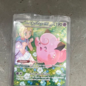 Lillie's Clefairy EX - Samlarkort med Lillie och Clefairy i färgglad anime-stil. Perfekt för Pokémon-fans som vill utöka sin samling med en unik och charmig EX-kort. Kortet är i skyddsplast och har coola attacker som Fairy Zone och Full Moon Rondo.