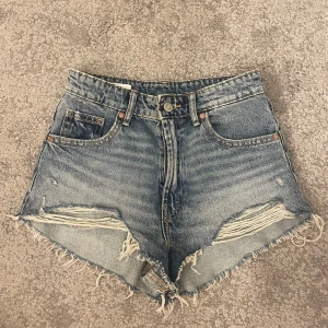 Jeansshorts från Zara - Snygga blå jeansshorts från Zara med råa, fransiga kanter. Shortsen har slitna detaljer framtill och klassisk femficksdesign. Perfekta för sommaren och ger en cool, avslappnad vibe. Shortsen är provade men aldrig använda då de är för små
