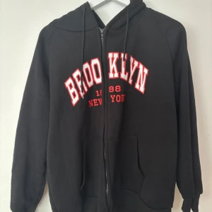 Svart Brooklyn hoodie med dragkedja - Svart hoodie med huva och dragkedja framtill. Stor BROOKLYN 1998 NEW YORK-tryck i vitt och rött över bröstet. Mjuk bomullskänsla, långa ärmar och kängurufickor. Perfekt för chill dagar eller streetstyle.