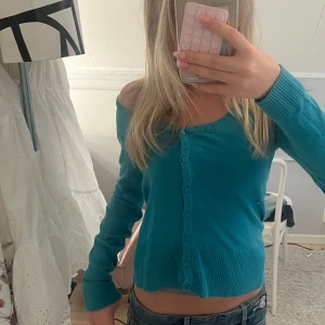 Turkos kofta med knappar - Säljer en turkos kofta med långa ärmar och knappar hela vägen framtill. Modellen är croppad och har breda ribbade muddar vid ärmslut och nederkant. Perfekt att bära öppen eller stängd för olika looks.
