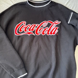 Svart Coca-Cola sweatshirt med tryck - Svart sweatshirt med stort Coca-Cola-tryck i rött och vitt på bröstet. Tröjan har vita ränder på krage och ärmslut, samt lång ärm. Perfekt för dig som gillar streetwear och vill sticka ut med en ikonisk logga.