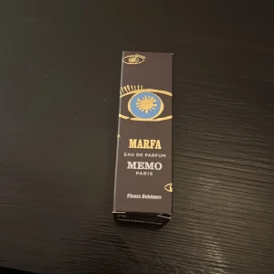 Memo Paris Marfa Eau de Parfum - Parfymen Marfa från Memo Paris i en elegant svart kartong med guld- och blått ögonmotiv. Texten är i guld och vitt, och förpackningen har en lyxig och konstnärlig känsla. Doften tillhör serien Fleurs Bohèmes.