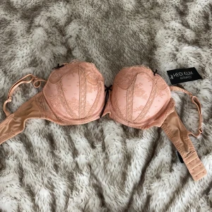 Ny  BH från Heidi Klum - Superfin Bh i persikofärgad spets från Heidi Klum Intimates. Detaljerad med broderad spets, små svarta rosetter och vadderade kupor. Snygg balconette-modell med justerbara axelband och hakknäppning bak. 