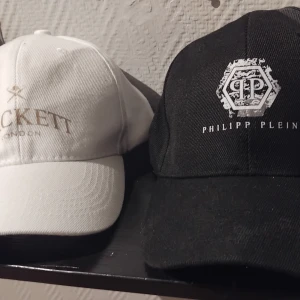Svart och vit keps Philipp Plein & Hackett - Två stilrena kepsar: en svart från Philipp Plein med vit logga framtill och en vit från Hackett London med broderad text i guld. Båda har klassisk böjd skärm och justerbar passform. Perfekta accessoarer för en clean och sportig look.