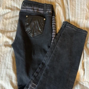 Svarta jeans med skinn och dragkedjor - Unika svarta jeans med gråa ränder, detaljer i fejkskinn på fickor och lår samt flera dragkedjor på ena benet. Snygg mix av material och cool streetstil. Perfekta för dig som vill sticka ut med din outfit.