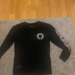 Svart långärmad Chrome Hearts tröja - Svart långärmad tröja från Chrome Hearts med vita tryck på bröstet, ryggen och båda ärmarna. Snyggt grafiskt mönster längs ärmarna och klassisk logga på bröstet. Perfekt för dig som gillar streetwear och vill sticka ut.