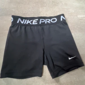 Svarta Nike Pro shorts - Svarta Nike Pro shorts med bred resår i midjan där det står NIKE PRO i vitt. Tight passform och korta ben, perfekt för träning. Materialet är stretchigt och andas, med en liten vit Nike-logga på ena benet.