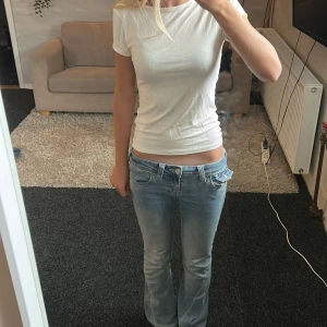 Vit t-shirt med rosett i satin - Supersöt vit t-shirt i mjukt material med korta ärmar och en snygg v-ringning i ryggen som pryds av en stor satinrosett. Perfekt för dig som gillar enkla plagg med en twist. Sitter figurnära och är lätt att matcha till jeans eller kjol.