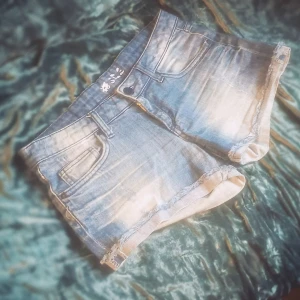 Blå jeansshorts med fransig kant - Snygga blå jeansshorts från B. Style med slitna detaljer och fransig kant nertill. Klassisk femficksmodell med knapp och dragkedja framtill. Perfekta för varma dagar när du vill ha en avslappnad och cool look.