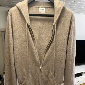 Beige hoodie i kashmir från John Henric - Mjuk och lyxig beige hoodie från John Henric i 100% kashmir. Tröjan har dragkedja framtill, huva med dragsko och två fickor. Perfekt för dig som gillar en clean och chill stil med premiumkänsla.