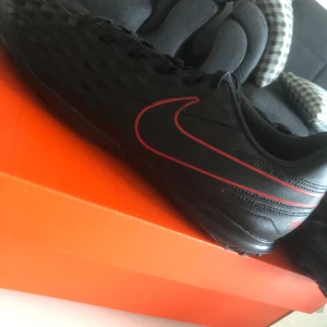 Svarta Nike fotbollsskor med röd swoosh - Svarta fotbollsskor från Nike med en snygg röd swoosh längs sidan. Skorna har snörning och en texturerad ovandel för bättre bollkontroll. Perfekta för dig som vill sticka ut på planen med stilrena detaljer.