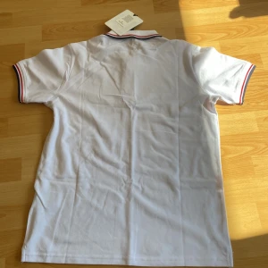 Vit Moncler pikétröja med ränder - Vit pikétröja från Moncler med klassisk krage och knappar framtill. Tröjan har Moncler-logga på bröstet och röda samt blå ränder på krage och ärmslut. Tillverkad i bomull och har en clean, sportig vibe som passar perfekt till jeans eller shorts.