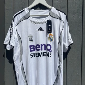 Real Madrid Ronaldo tröja Adidas XL - Real Madrid fotbollströja med Ronaldo 9 på ryggen, Adidas-märke och Champions League-märke på ärmen. Vit med svarta detaljer och lila BenQ Siemens-tryck. Broderat klubbmärke och FIFA Award-märke på bröstet. Tillverkad i lätt och ventilerande material.