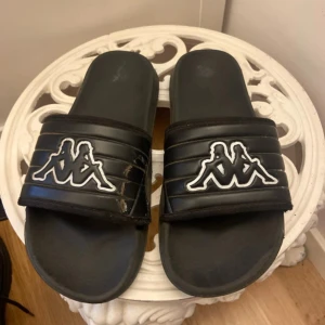 Svarta Kappa sandaler med logga - Svarta sandaler från Kappa med den klassiska vita loggan framtill. Skorna har en bred rem över foten med vadderad känsla och mönstrad sula för extra grepp. Perfekta för sommaren och chill dagar.