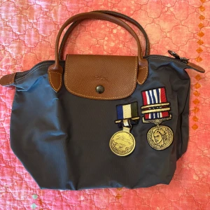 Longchamp handväska marinblå - Snygg marinblå handväska från Longchamp med bruna skinnhandtag och lock. Väskan har dragkedja och tryckknapp samt två coola medaljpatches framtill. Klassisk och rymlig modell i slitstarkt syntetmaterial med detaljer i präglat skinn.