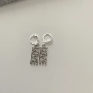 Silvriga örhängen med LOVE-text - Snygga silvriga örhängen med hängande rektangulära berlocker där det står LOVE i glittriga stenar. Bokstäverna är täckta av små stenar som ger extra bling. Perfekta för dig som vill ha ett statement-smycke med cool vibe.