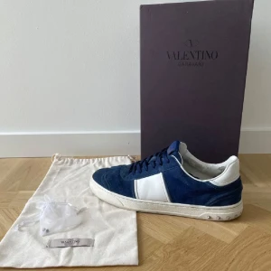 Valentino Garavani blå sneakers mocka - Snygga sneakers från Valentino Garavani i blå mocka med vita skinnpaneler och coola nitar på hälen. Skorna har rund tå, snörning och en platt sula. Perfekta för dig som vill ha en lyxig och trendig look med streetkänsla.