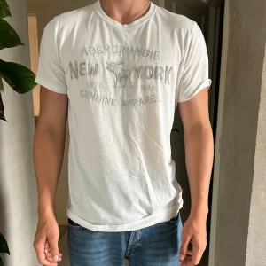Vit Abercrombie & Fitch t-shirt M - Vit t-shirt från Abercrombie & Fitch med tryck på bröstet och klassisk rund hals. Skön passform och tillverkad i mjuk bomull. Perfekt basic-plagg med diskret logga och stilren look.