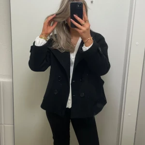 Svart skepparkavaj med dubbelknäppning - Snygg svart skepparkavaj med klassisk dubbelknäppning och bred krage. Jackan har en rak passform och är perfekt för dig som gillar en stilren look. Passar till både jeans och kostymbyxor. Materialet känns mjukt och värmande.