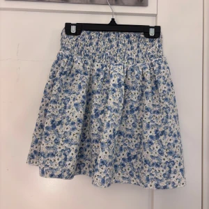 Blommig kortkjol med smock i midjan - Supersöt kortkjol med blått och vitt blommönster och smockad midja. Står ej storlek men skulle säga 158/164 eller XS 💕bra skick