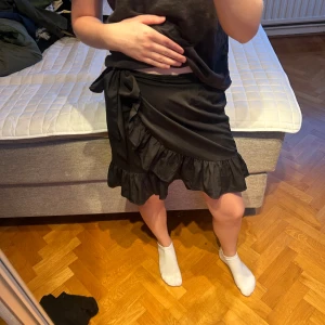 Svart omlottkjol med volang - Supersnygg svart omlottkjol med volangkant och knyt i midjan. 